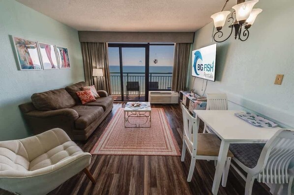 Un salon comprenant un canapé, un fauteuil, une table basse et une table à manger. Un téléviseur est fixé au mur, et un balcon offre une vue sur l’océan.