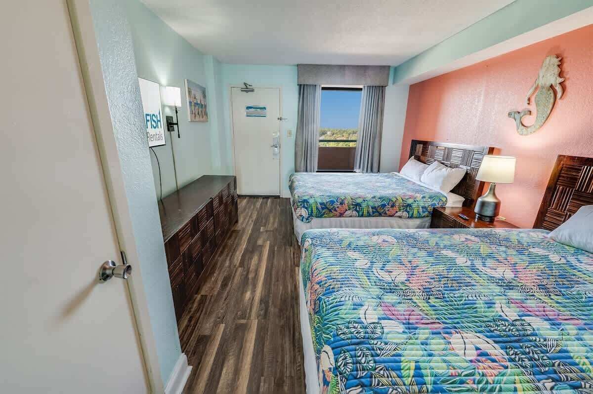 Habitación de hotel con dos camas, un escritorio, una silla, una puerta y vistas al exterior.