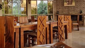 A dining area with wooden furniture, large windows, and a stone wall with a mounted picture. - Kambi Mstuni Resort (Ndaragwa)