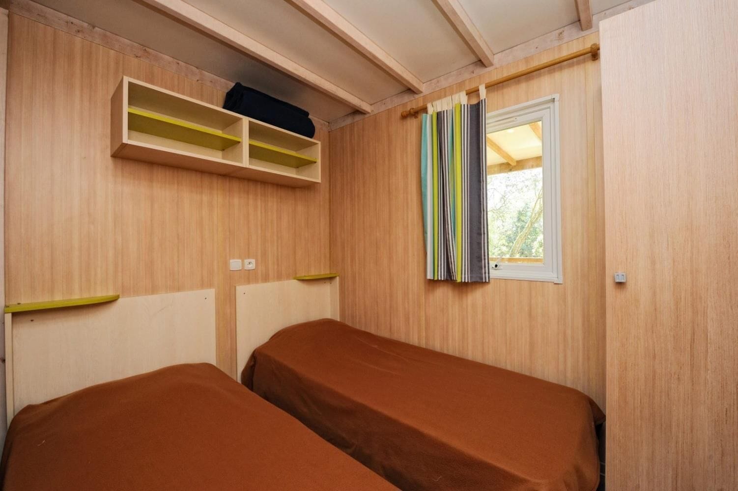 Une petite pièce avec deux lits, des murs en bois, une fenêtre avec des rideaux à rayures et une étagère au-dessus des lits.