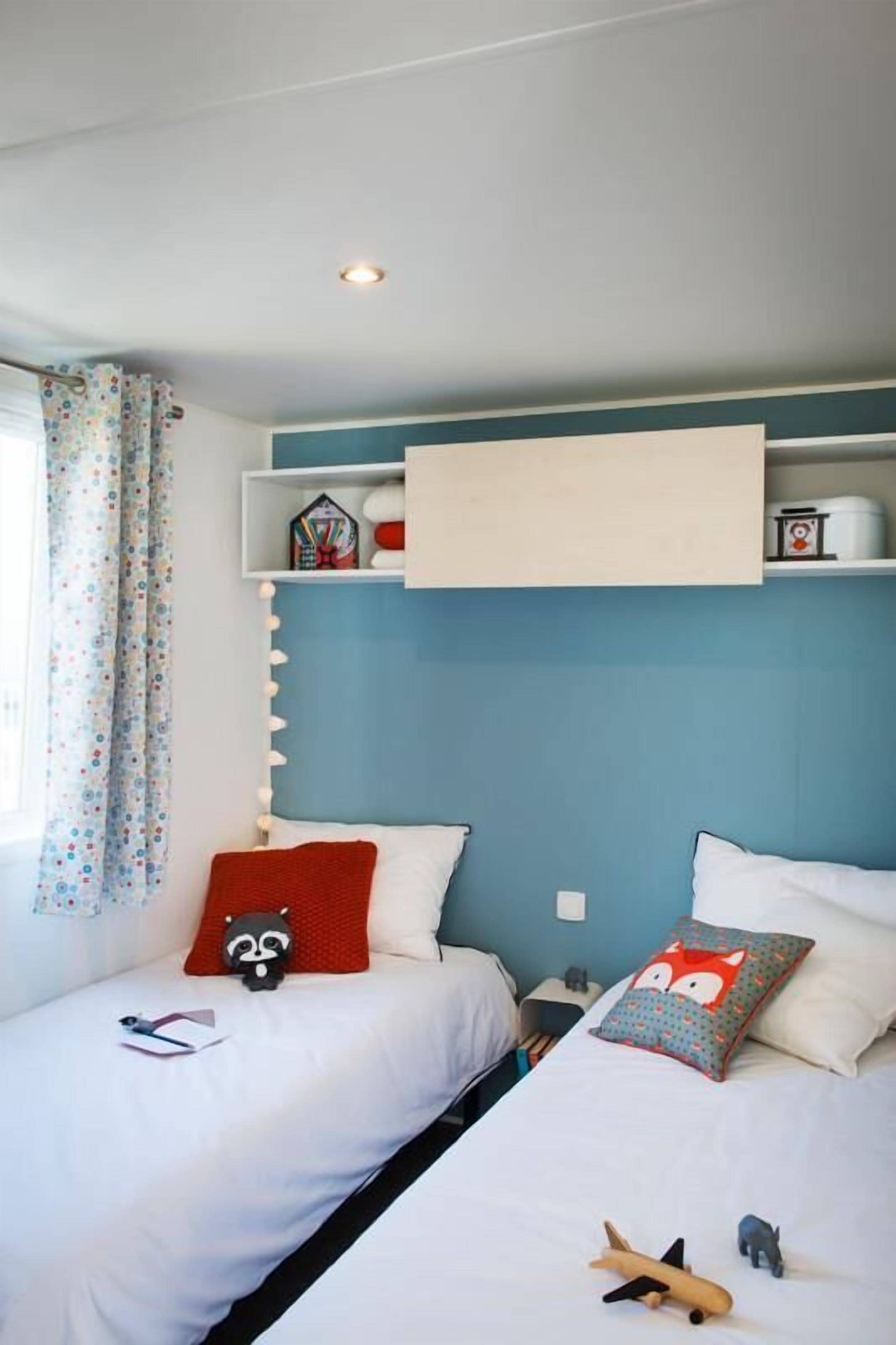 A bedroom with two beds, a shelf with toys, and a window with curtains.