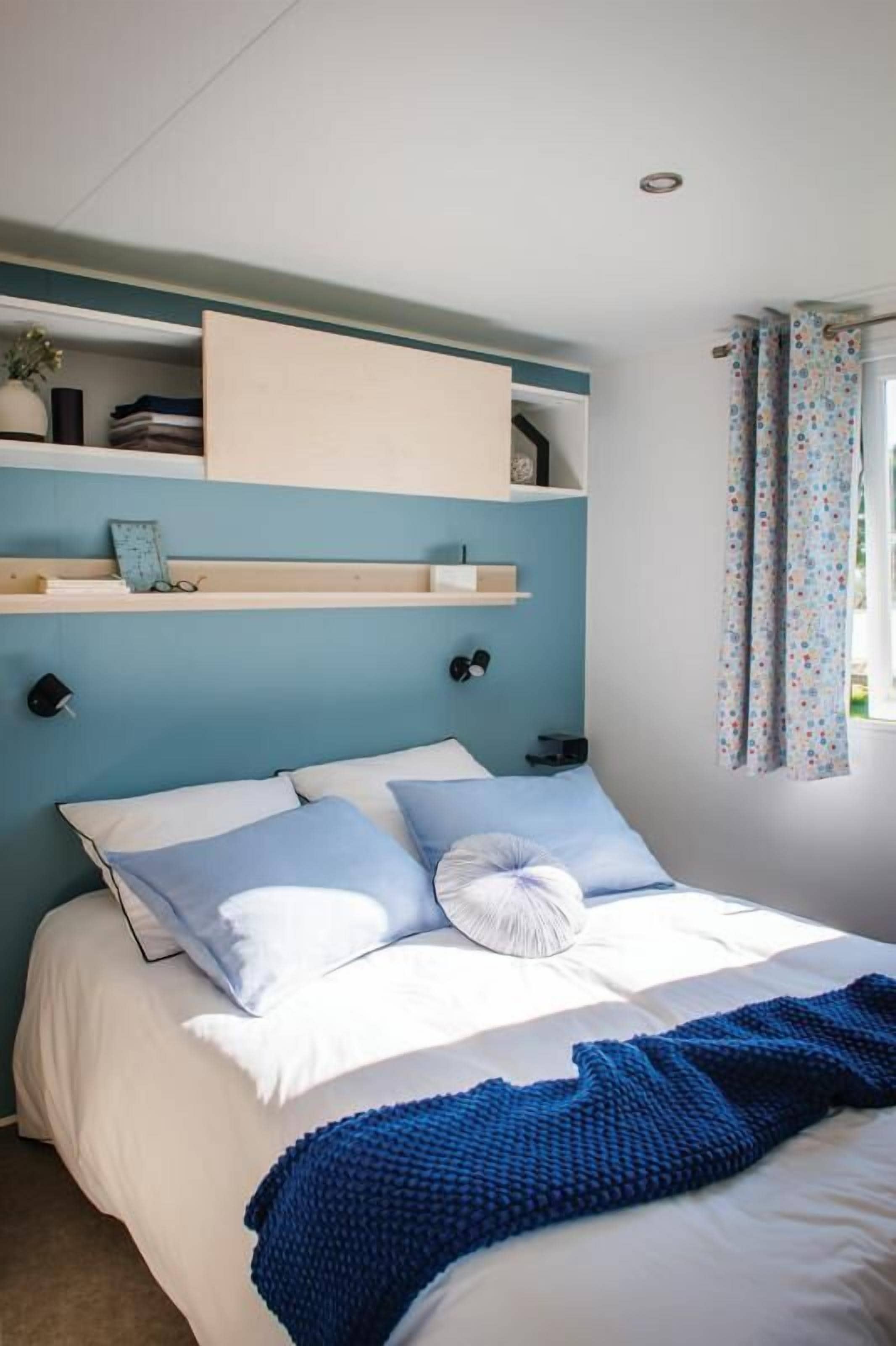 A bedroom with a bed, pillows, a blue blanket, and a shelf with books and a clock.