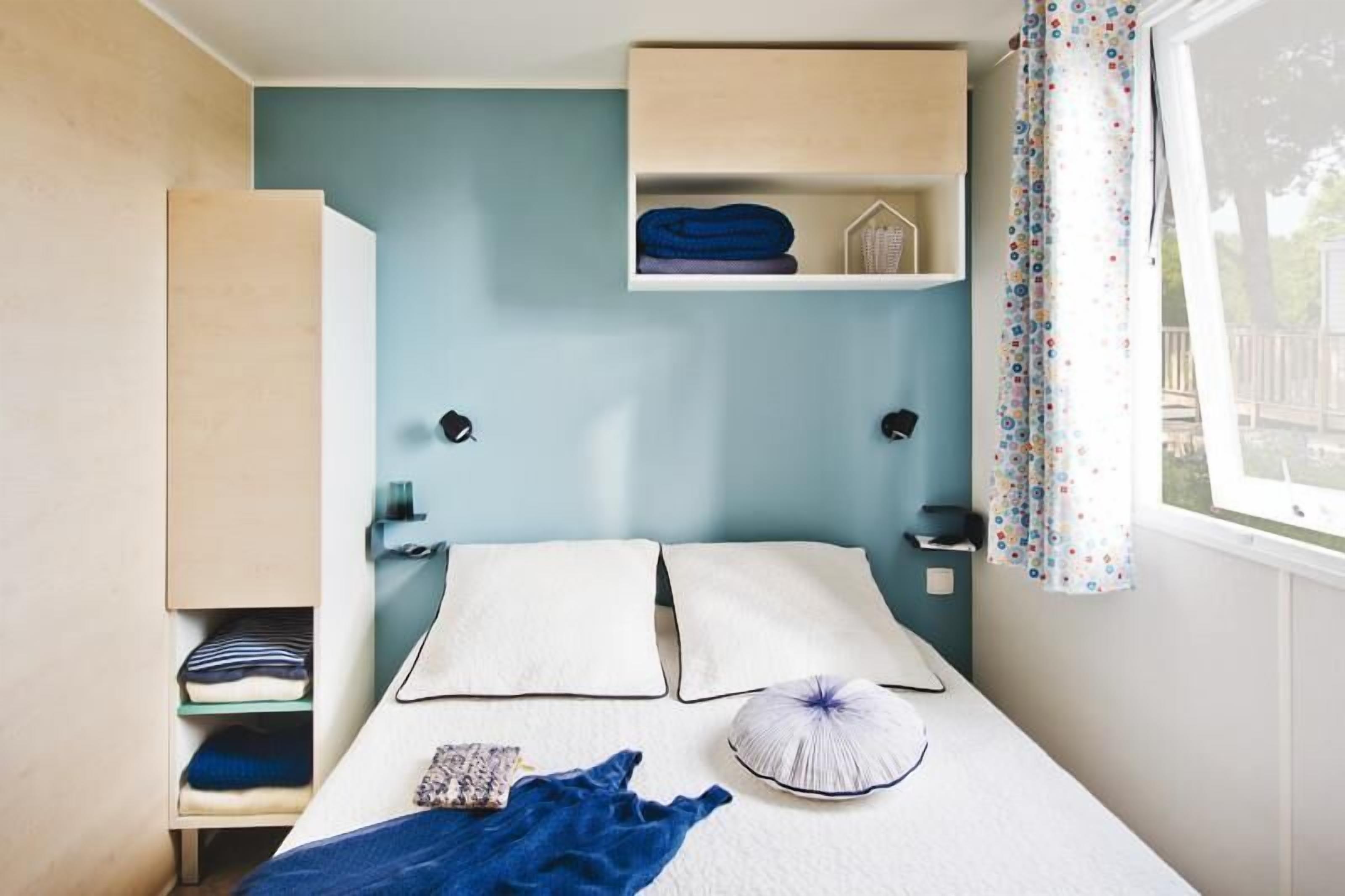 A compact bedroom with a bed, pillows, a blue blanket, a small shelf with folded clothes, and a window with curtains.