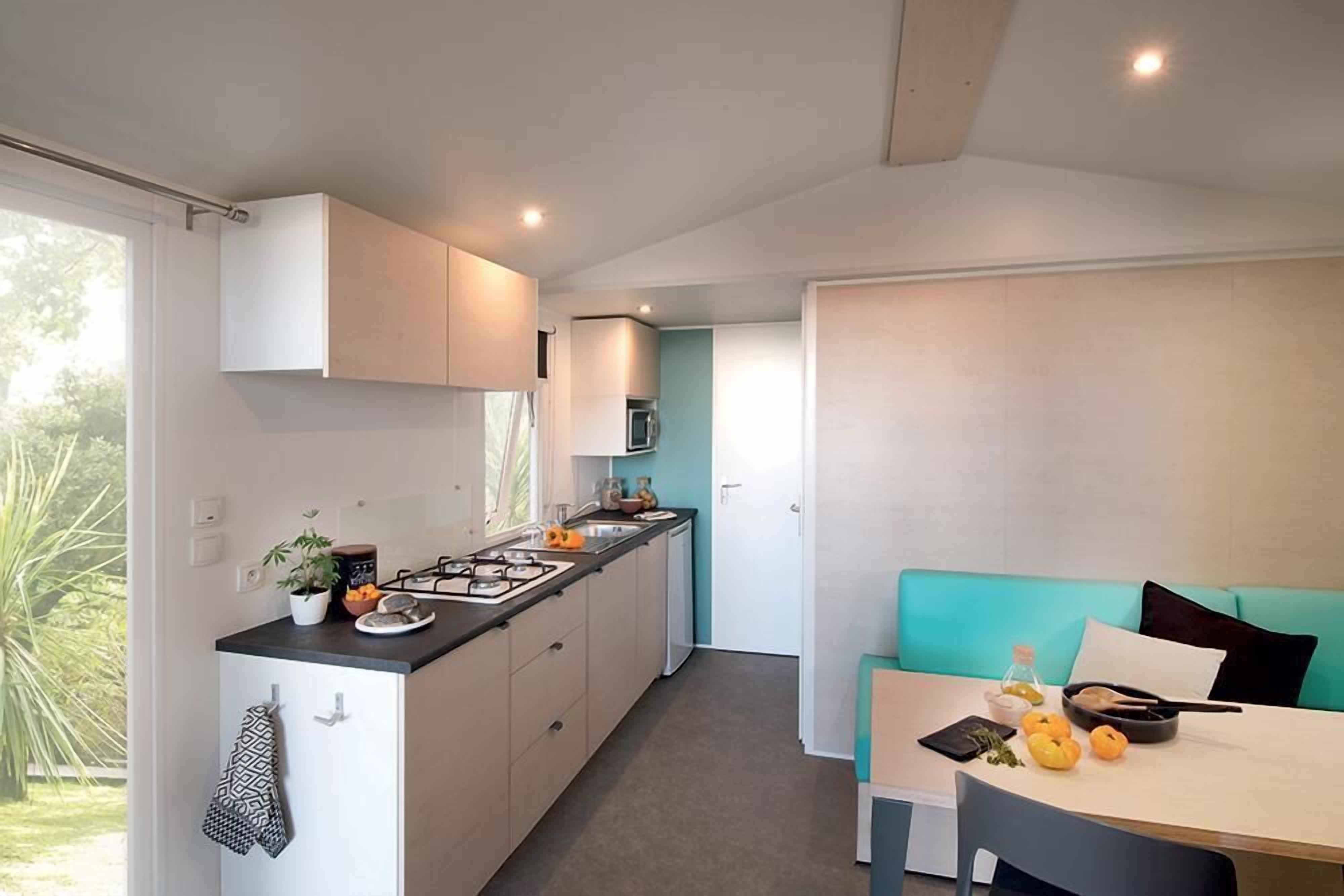 A compact kitchen with a central island, a dining area with a teal sofa, and a view of greenery outside.