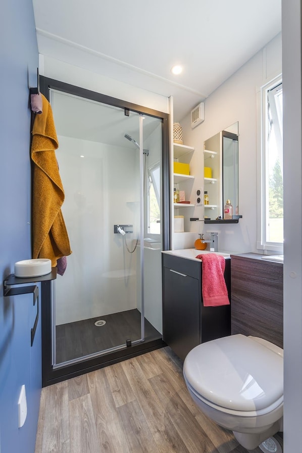 A modern bathroom with a glass shower enclosure, a toilet, a sink with a mirror, and a shelf with toiletries. - Camping Campo di Liccia (Bonifacio)