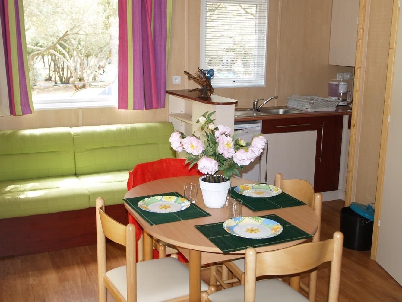 A dining area with a wooden table set for four, a green sofa, and a window with curtains.