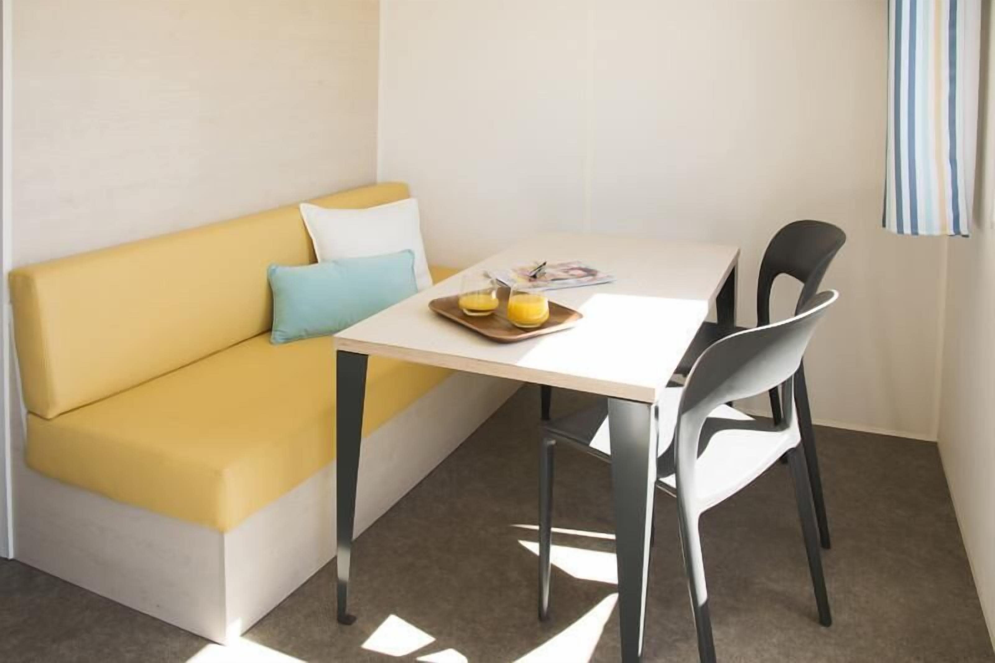 Une petite pièce avec un canapé jaune, une table blanche et deux chaises noires. Sur la table, on trouve un plateau avec deux verres de jus d’orange.