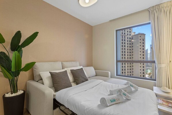 A hotel room with a bed, a large window with a city view, a potted plant, and a nightstand with a lamp.