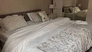 Un llit ben fet amb un edredó blanc i gris amb dibuixos, un panell de fusta al capçal i una taula de nit amb una làmpada i objectes decoratius.