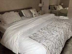 Una cama bien hecha con colcha blanca y gris estampada, cabecera de madera y mesita de noche con lámpara y objetos decorativos.