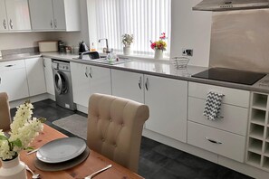 A modern kitchen with white cabinetry, a built-in washing machine, a dining area with a table and chairs, and a window with blinds. - Beach House Apartment, Sleeps 4 With Patio (Southport)