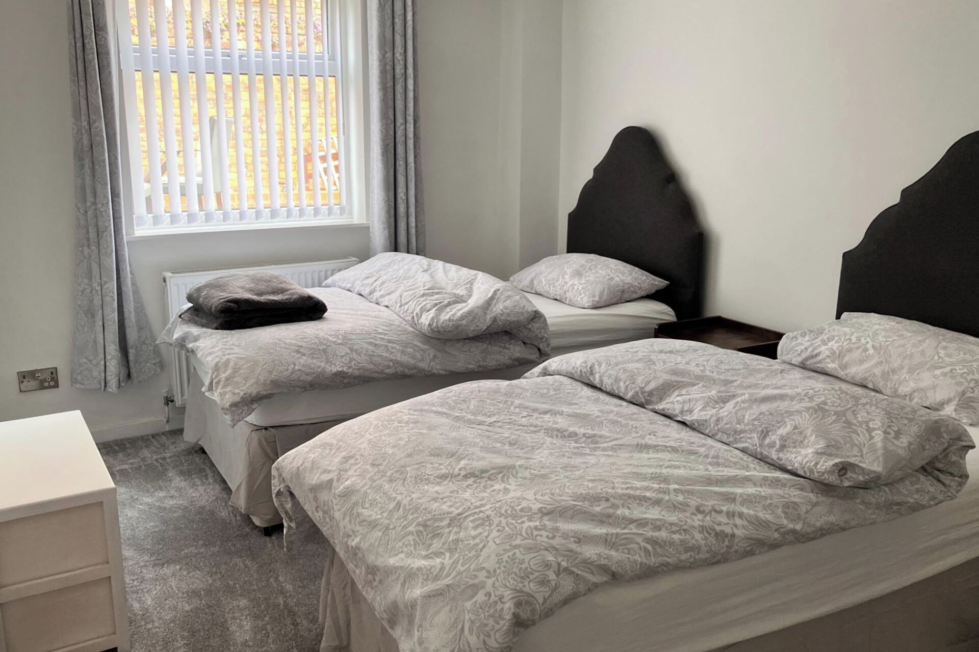 A bedroom with two beds, a window with blinds, a bedside table, and a dresser.
