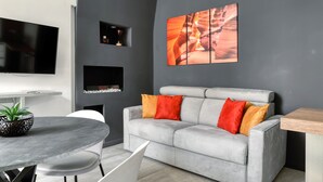 A modern living room with a grey sofa, two red and one yellow cushion, a round dining table with chairs, a potted plant, and a flat-screen TV mounted on the wall. - Farini 74 Design Apartment – Metro M3, WiFi, AC (Milan)