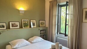 A neatly made bed with white linens and towels, a bookshelf with books, a wall-mounted light, and a window with curtains.