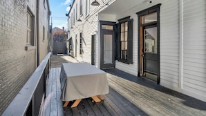 Ein schmaler Gang mit einem Tisch, der mit einer Tischdecke bedeckt ist, einer Holzterrasse und einem Gebäude mit weißer Schalung und schwarzen Fensterläden.
