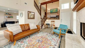 A spacious living room with a brown leather sofa, a colorful rug, a stone fireplace, and a staircase leading to an upper level. - Second Wind Bay House - Fish, Float, Fun! (Freeport)