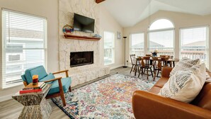 A living room with a stone fireplace, a television mounted above it, a dining area with a table and chairs, and large windows with blinds. - Second Wind Bay House - Fish, Float, Fun! (Freeport)