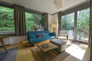 A modern living room with a blue sofa, a side table, a lamp, and a view of a forest through large windows.