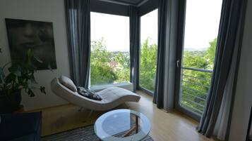 Ein modernes Wohnzimmer mit einer Couch, einem Glastisch und Blick auf eine grüne Landschaft durch große Fenster.