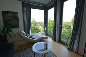A modern living room with a sofa, a glass coffee table, and a view of a green landscape through large windows.