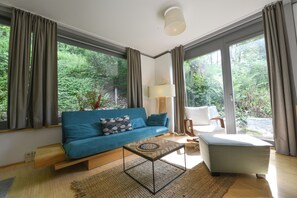 A modern living room with a blue sofa, a small coffee table, and a chair. Large windows offer a view of greenery outside.