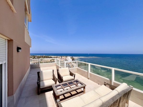 A balcony with outdoor furniture, a clear view of the ocean, and a building facade.