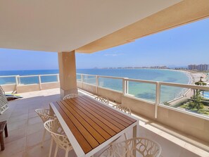 A balcony with a wooden table and white chairs, overlooking a beach and cityscape.