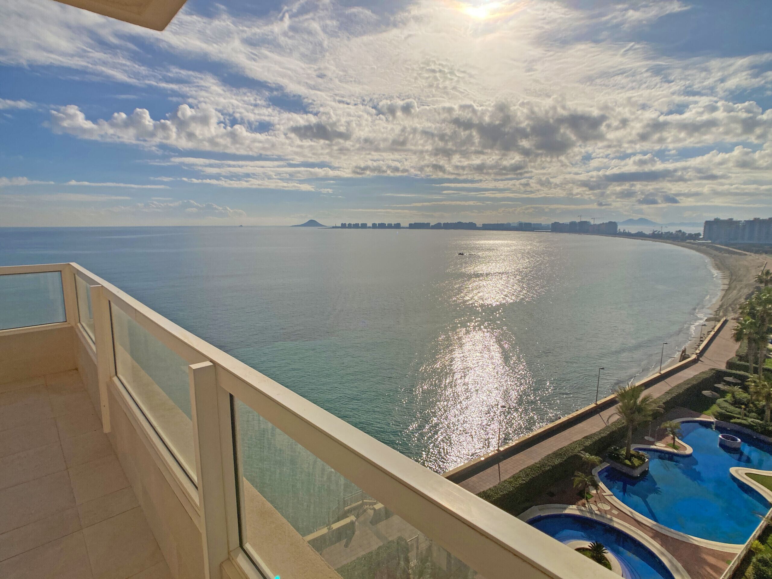 A balcony with a view of a calm sea, a beach, and a swimming pool.