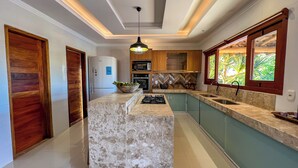A modern kitchen with a central island, built-in appliances, and a view of the outdoors through a window. - Pousada Refúgio Verde Trancoso (Porto Seguro)