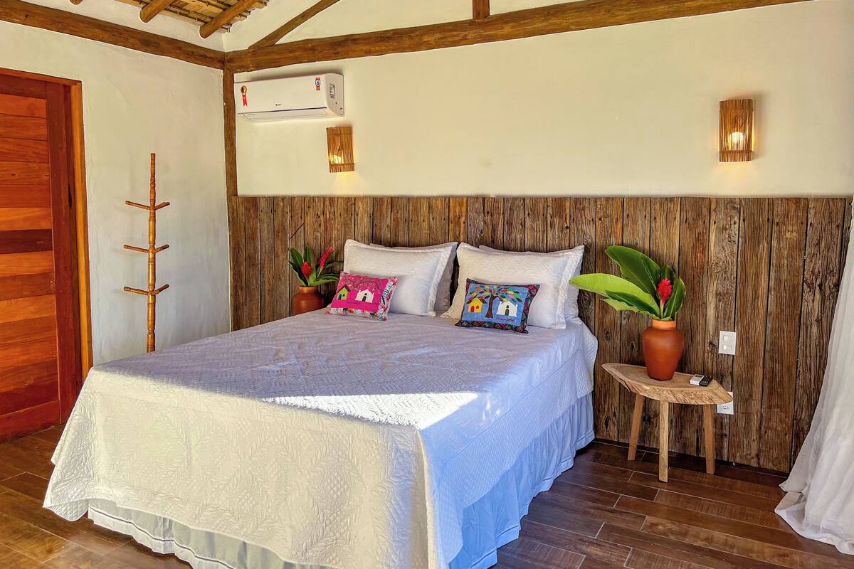 Une chambre à coucher avec une tête de lit en bois, un lit avec une literie blanche, une table de chevet avec un vase et une plante en pot.