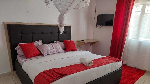 A neatly made bed with a red and white color scheme, a bedside table, a TV mounted on the wall, and a red curtain. - White House Lounge & Accommodation  (Eldoret)