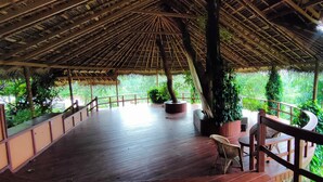 A covered wooden deck with a thatched roof, surrounded by lush greenery. - HORTUS MALABARICUS RESORT (Talappilly)