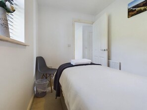 Una habitación pequeña y ordenada con una cama individual, una silla, un radiador y una puerta que da a otra habitación.