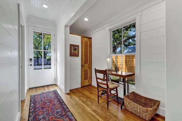A hallway with a patterned rug, a wooden chair, a wicker basket, and a table with a lamp.