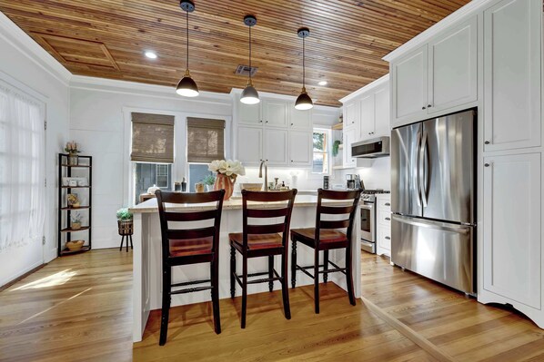 A modern kitchen with wooden flooring, white cabinets, a stainless steel refrigerator, and pendant lighting.