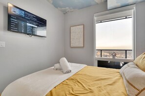 A hotel room with a bed, a TV mounted on the wall displaying a program guide, a framed artwork, and a large window offering a city view. - Legends - Executive Suite, Luxury Amenities, Pool (Edmonton)