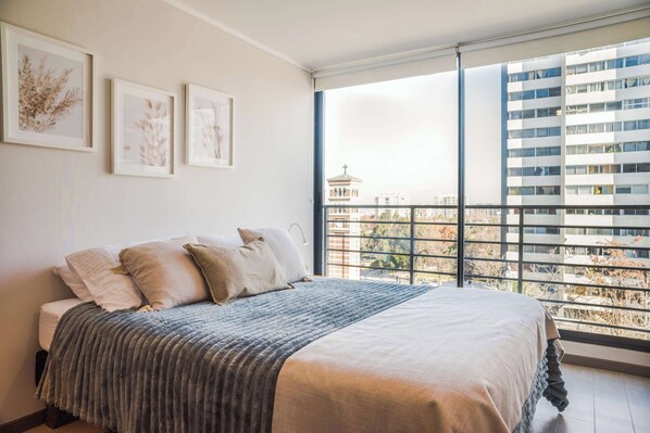 A bedroom with a large bed, a view of buildings through a window, and framed artwork on the wall.