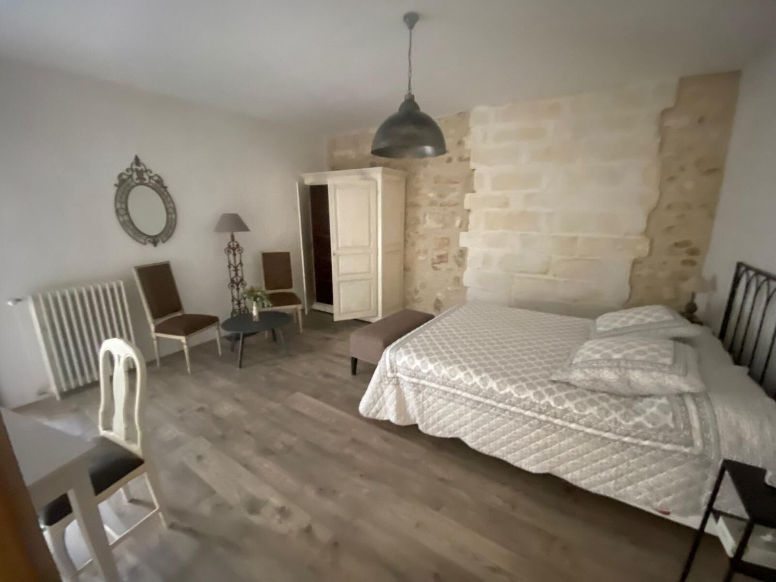 A bedroom with a large bed, a chair, a small table, and a lamp. There is a radiator on the left and a wardrobe against the wall.