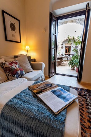 Een kamer met een bed, een zithoek met een boek, een lamp en uitzicht op een terras buiten met planten en meubilair.