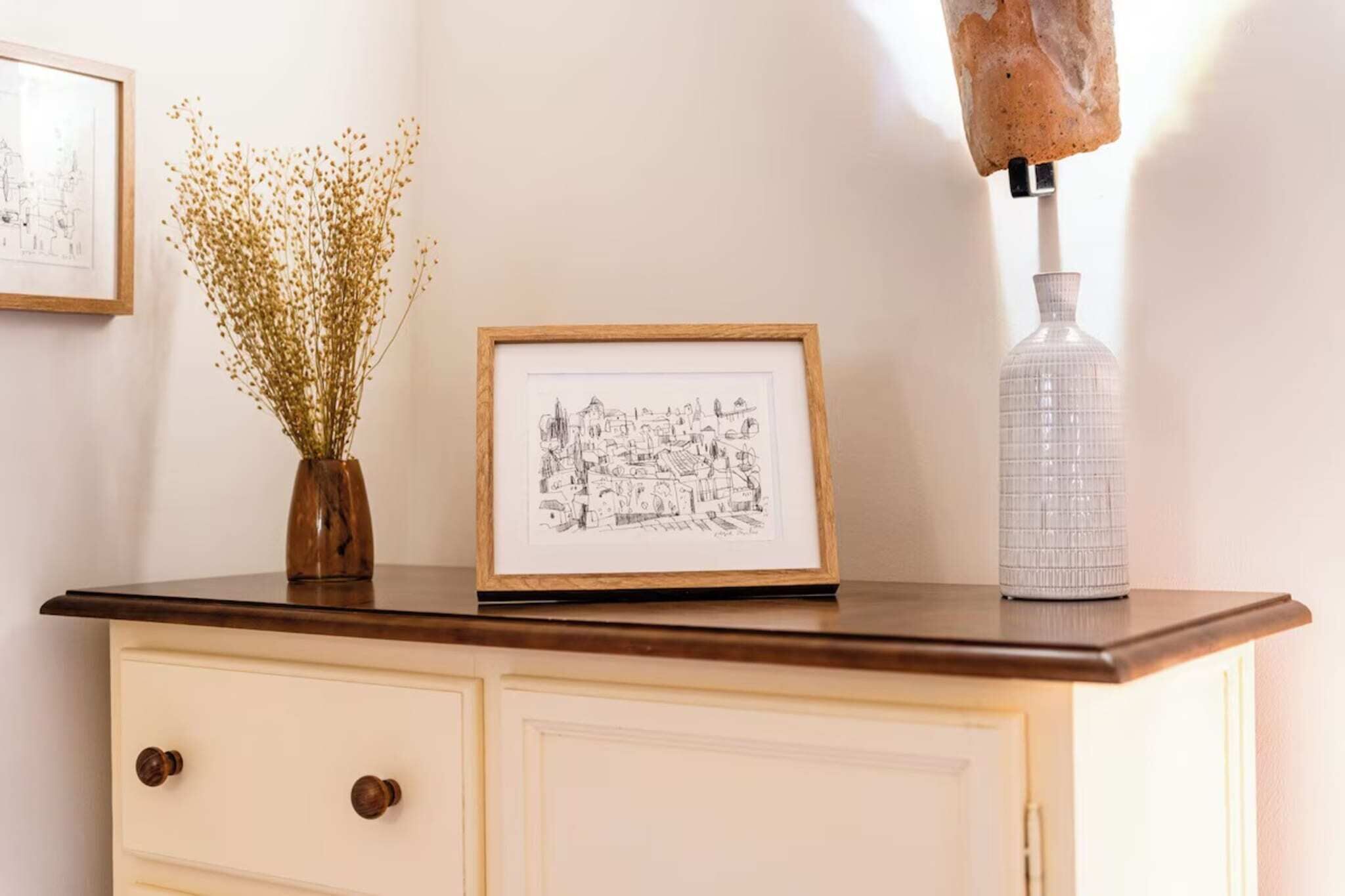 Une esquisse encadrée posée sur une commode, un vase avec des fleurs séchées et une bouteille transparente.