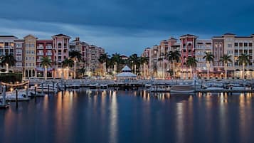 A marina with boats docked, a covered pavilion, and a row of multi-story buildings with balconies and palm trees.