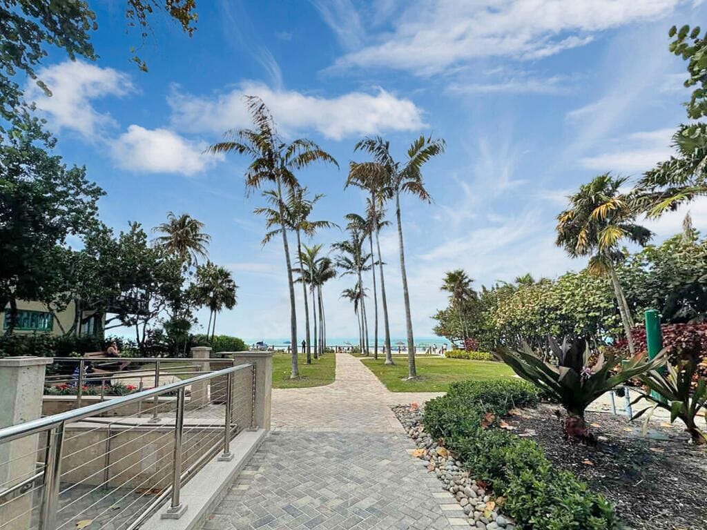 A pathway lined with palm trees leading to a beach, with a railing on one side and a landscaped area with plants on the other.