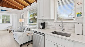 A modern kitchen with white cabinets, a dishwasher, and a sink. There is a window with blinds, a lamp, and a small table with a coffee maker. - Tropical Oasis for your Naples Getaway!  (Naples)