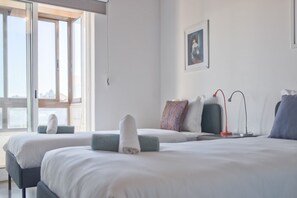 Ett hotellrum med två sängar, ett stort fönster, en tavla på väggen och ett skrivbord med en lampa.