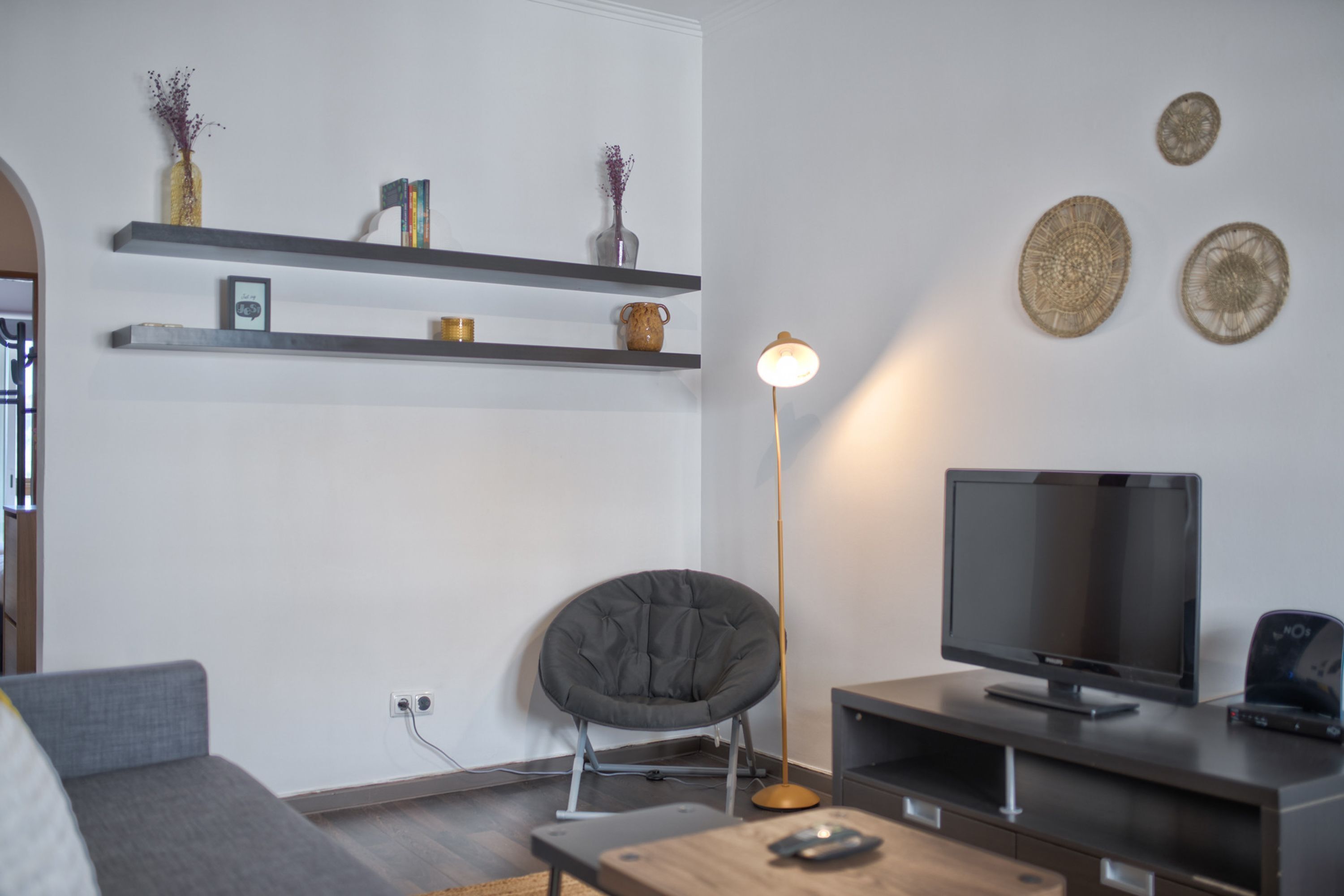 Ein modernes Wohnzimmer mit einem grauen Sofa, einem runden Sessel, einem Fernseher auf einem Schrank und Dekoration auf Regalen.