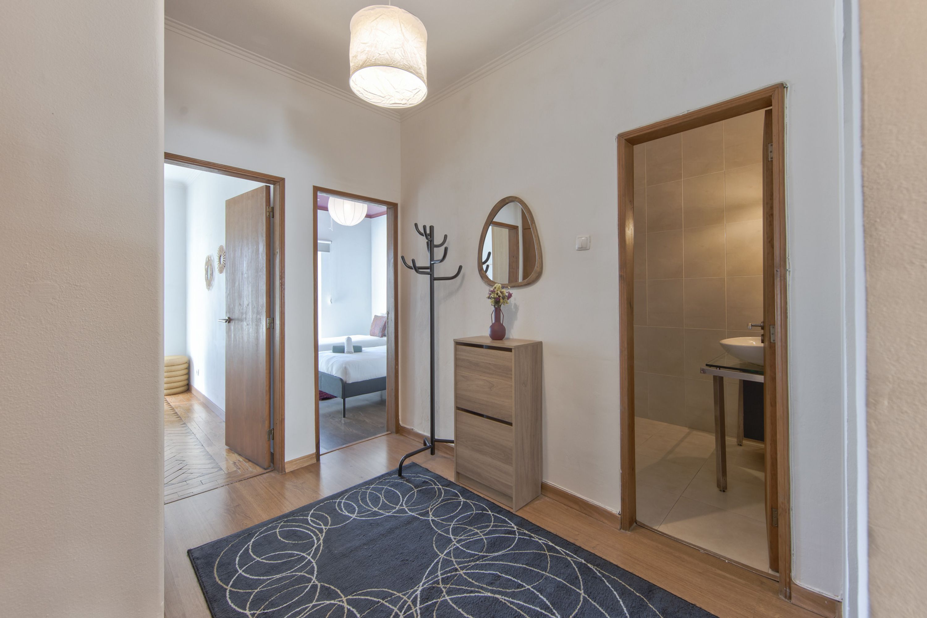A hallway with a wooden floor, a dark patterned rug, a coat rack, a mirror, and a door leading to a bathroom.