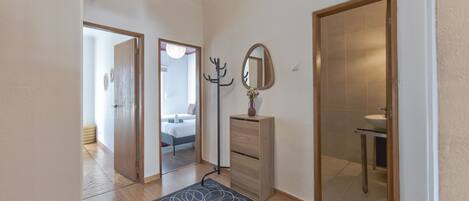 A hallway with a wooden floor, a dark patterned rug, a coat rack, a mirror, and a door leading to a bathroom.