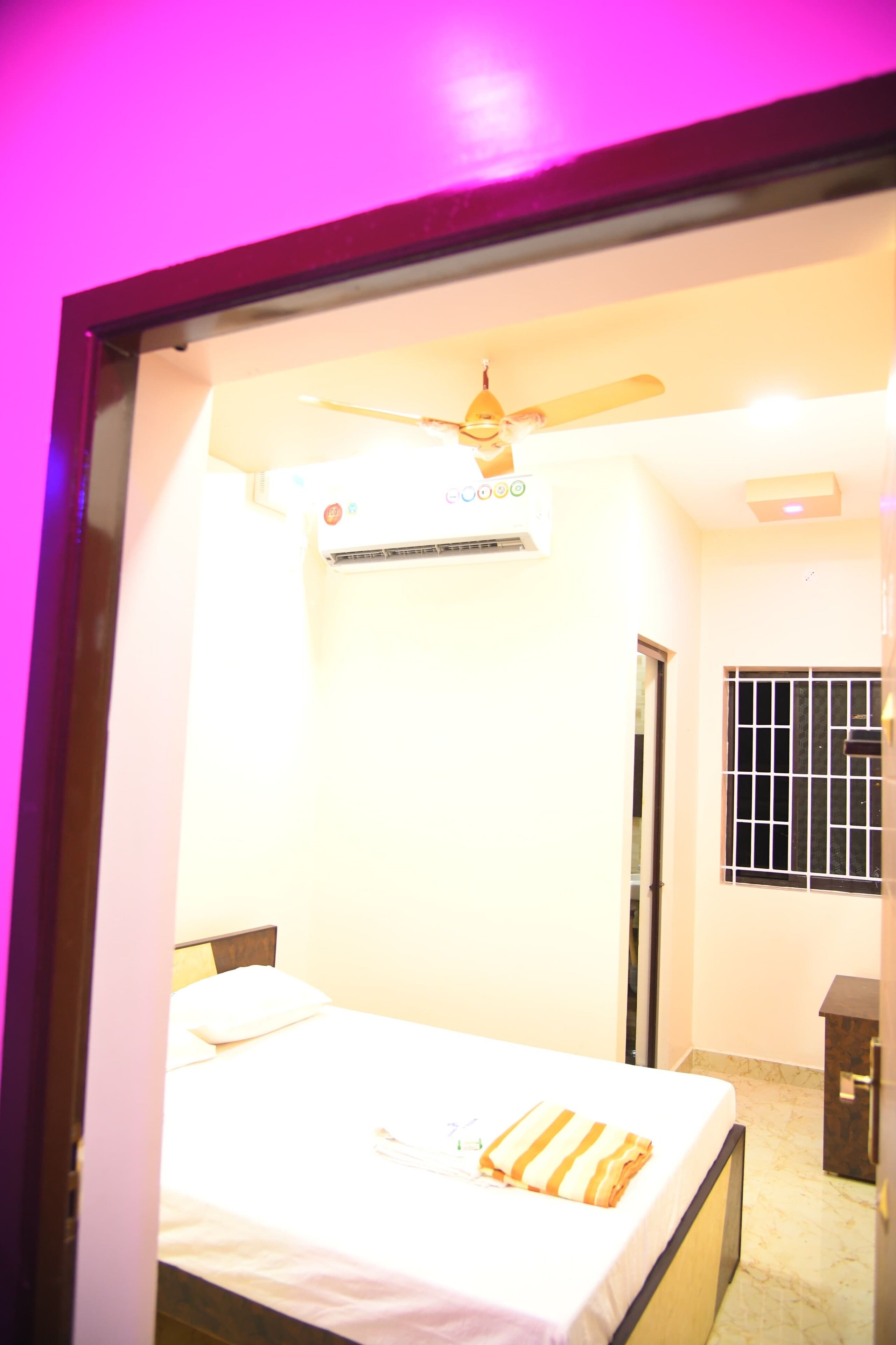 Un dormitorio con cama, unidad de aire acondicionado, ventilador de techo, ventana con rejas y una puerta.