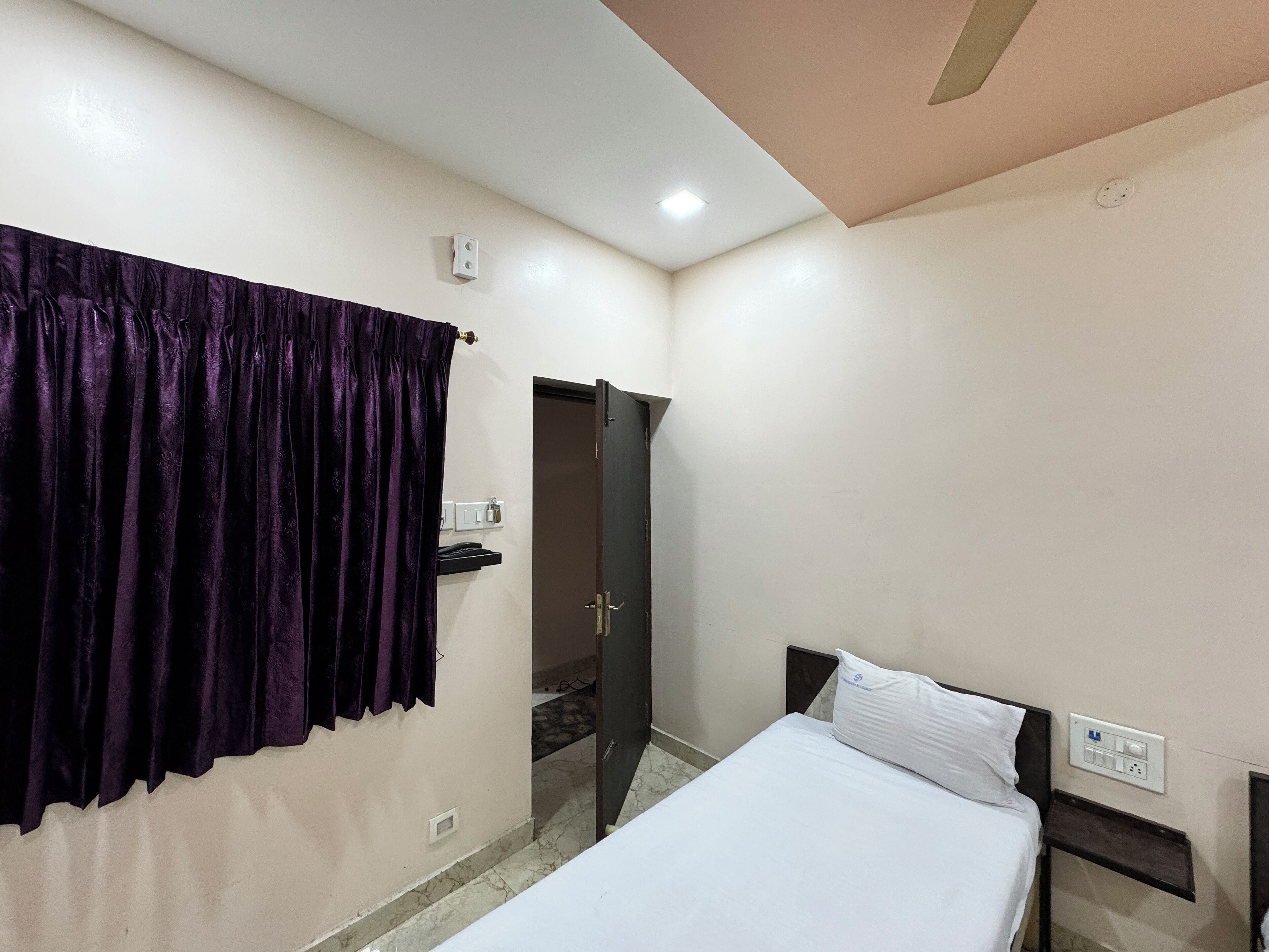 Een hotelkamer met een bed, een nachtkastje, een stopcontact en een deur die naar een andere kamer leidt.