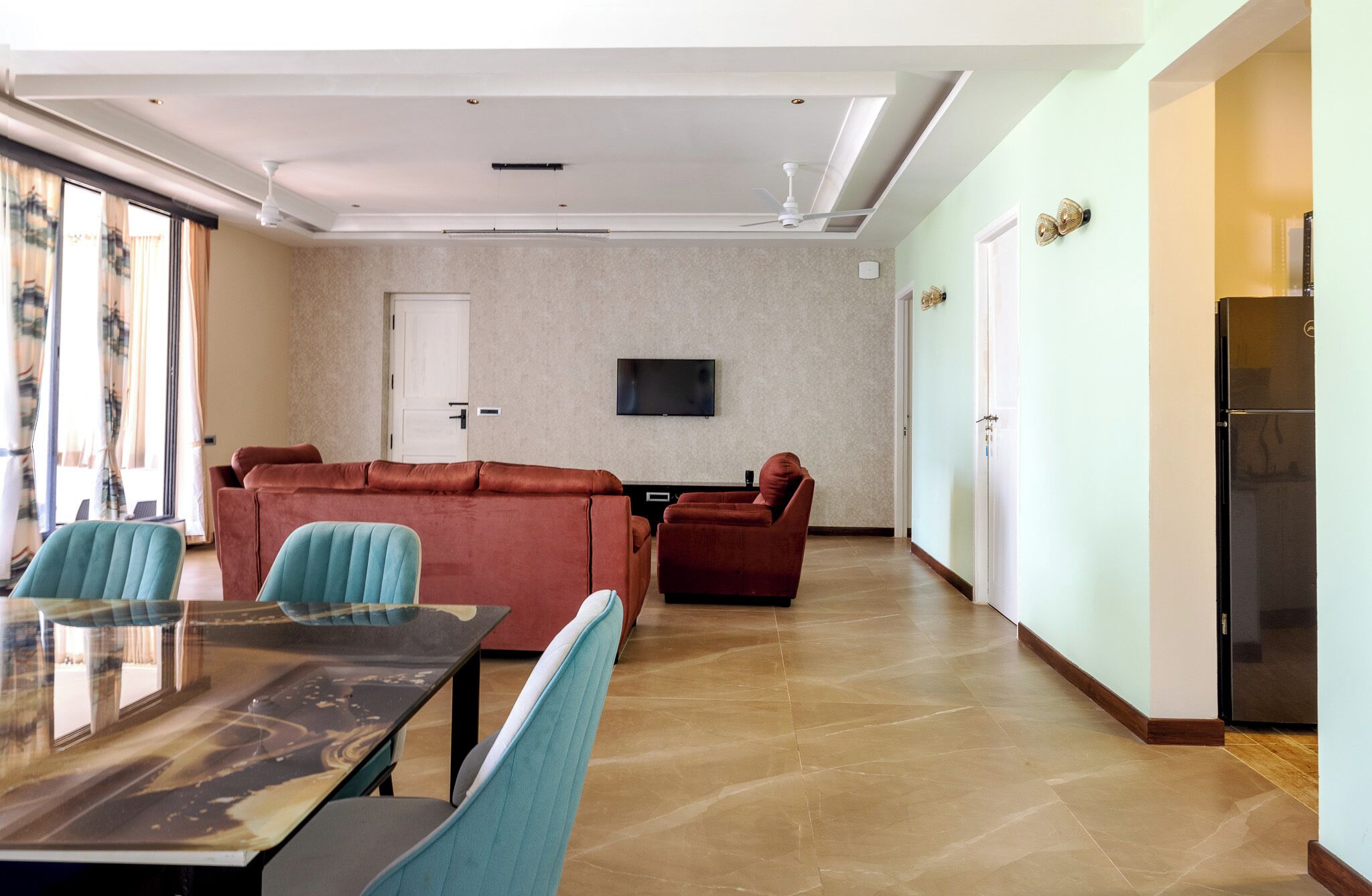 A spacious living room with a sofa set, a dining table with chairs, a flat-screen TV mounted on the wall, and a refrigerator.